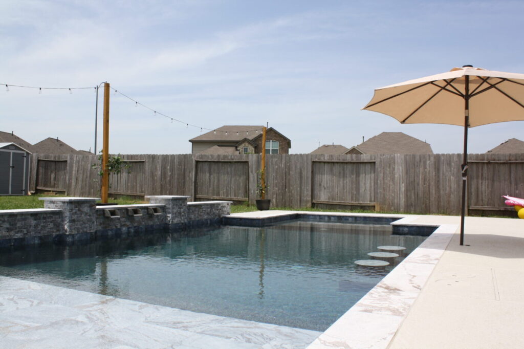 Texas Turf Tenders installed a geometric pool with a beach entry, tanning ledge, and in-water barstools. They added a raised spa with a rolled edge, a raised wall with columns and scuppers, plus surrounding turf and spray decking.