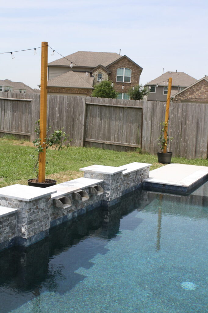 Texas Turf Tenders installed a geometric pool with a beach entry, tanning ledge, and in-water barstools. They added a raised spa with a rolled edge, a raised wall with columns and scuppers, plus surrounding turf and spray decking.
