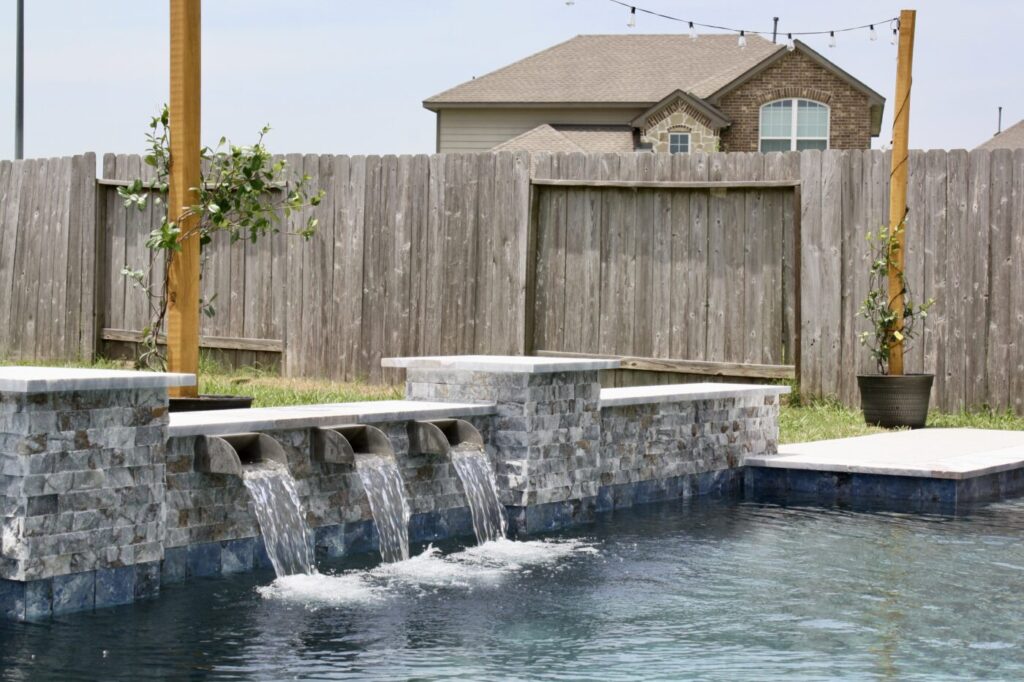 Texas Turf Tenders installed a geometric pool with a beach entry, tanning ledge, and in-water barstools. They added a raised spa with a rolled edge, a raised wall with columns and scuppers, plus surrounding turf and spray decking.
