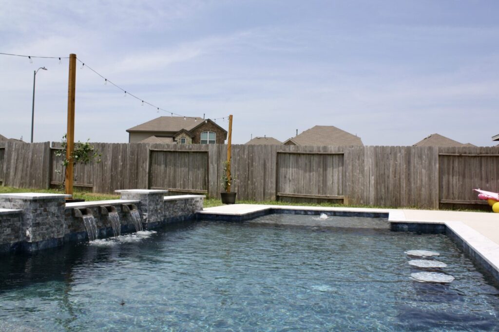 Texas Turf Tenders installed a geometric pool with a beach entry, tanning ledge, and in-water barstools. They added a raised spa with a rolled edge, a raised wall with columns and scuppers, plus surrounding turf and spray decking.