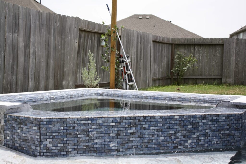 Texas Turf Tenders installed a geometric pool with a beach entry, tanning ledge, and in-water barstools. They added a raised spa with a rolled edge, a raised wall with columns and scuppers, plus surrounding turf and spray decking.